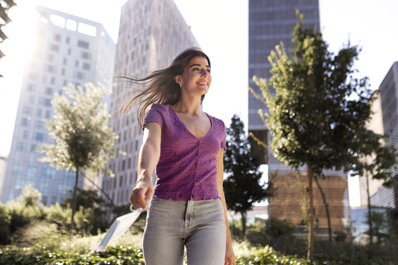 mujer sonriente entre edificios con mascarilla en mano